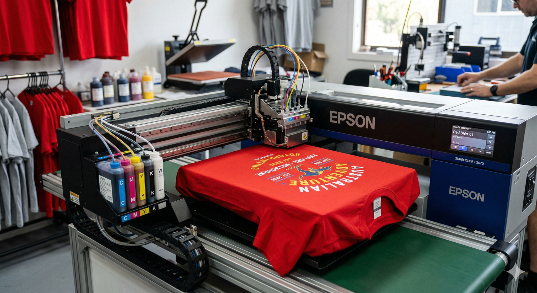 A high-end Epson SureColor F3070 direct-to-garment printer printing a detailed 'AUSTRALIAN ADVENTURE' graphic onto a red t-shirt. The print head is moving over the shirt, which is coming out of the machine on a green platen conveyor. The background shows a print workshop with racks of red t-shirts, various inks, and a person working on another piece of equipment.