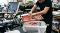 A close-up photograph of a printer manually screen printing a two-color logo onto a white t-shirt using a professional printing press. The logo features a red crest and dark blue typography. One screen with red ink is being applied with a squeegee, while a second screen for the dark blue details is visible on the carousel in a professional workshop setting